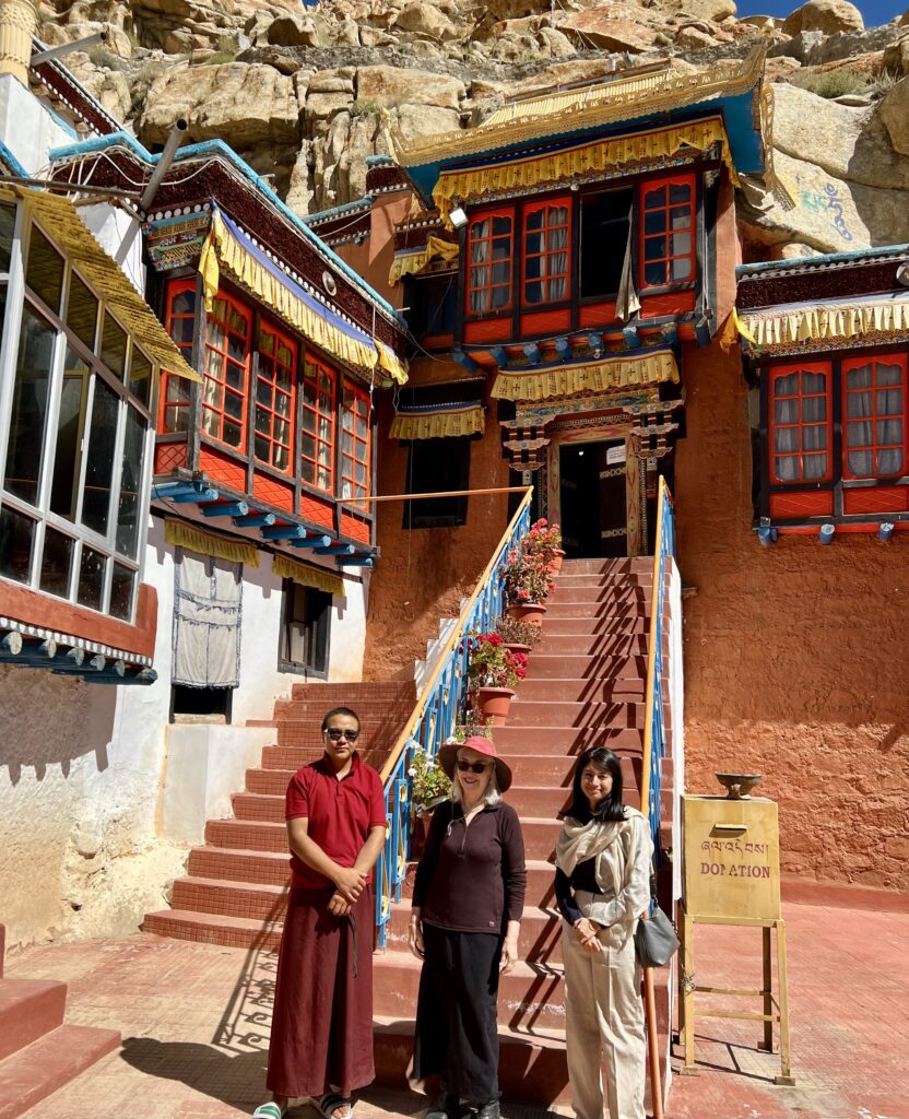 Three people stand near a staircase leading to a traditional Tibetan building.