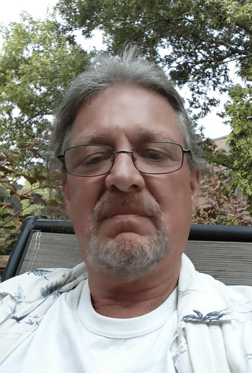 Middle-aged man with glasses outdoors, trees in background.