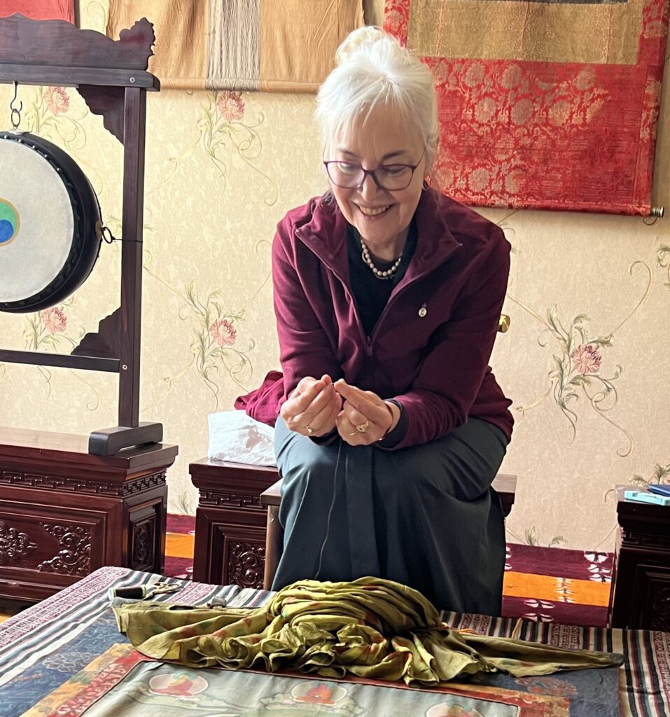 Elderly woman sitting at a wooden table with folded hands and a warm smile.