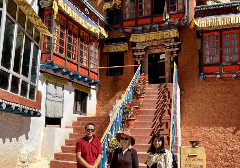 Three people stand near a staircase leading to a traditional Tibetan building.